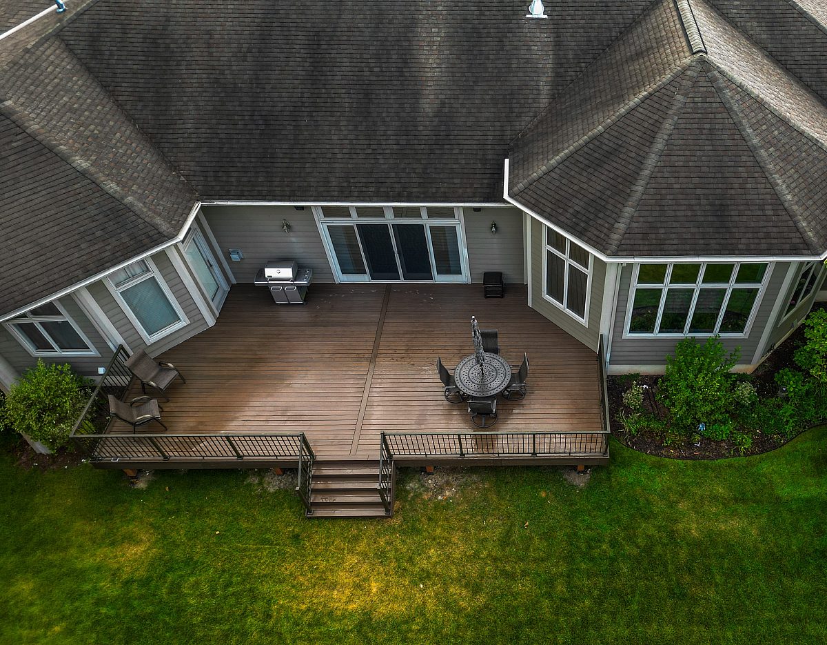 Amazing deck built by Green Shield Deck Builders seen from above with a drone. On the deck you can see a table, chairs, and umbrella. Near the house sits a shiny silver grill. Rich Tropical Walnut color deck boards and bronze aluminum baluster deck railing.