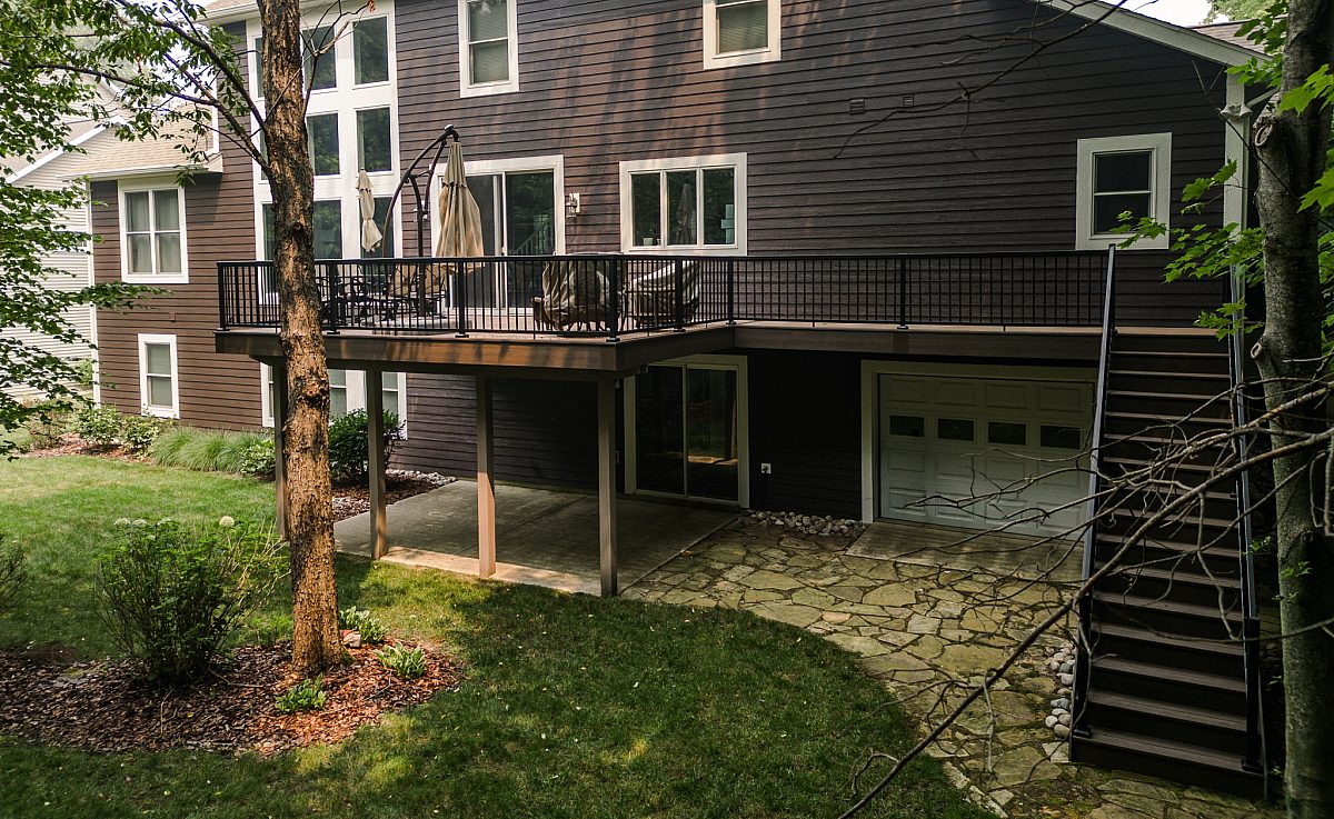 A sprawling Tigerwood composite deck with Tropical Walnut accents built by Green Shield Deck Builders on a brown home nestled in the woods. Project features black aluminum baluster and 14 standard steps leading to a grassy backyard.