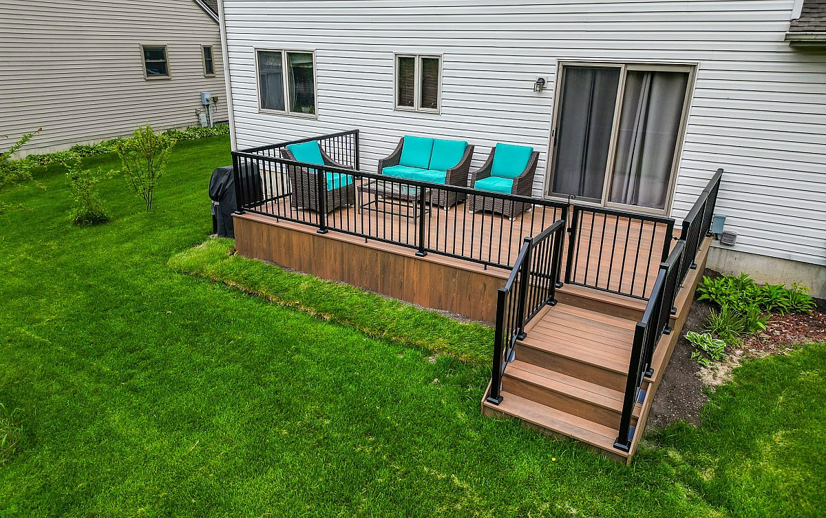 Green Shield deck with Tigerwood color composite deck boards and black aluminum baluster deck railing. Vibrant deck furniture seen on deck surface with glass coffee table. Image taken from front of deck showing green grass and white colored house siding.