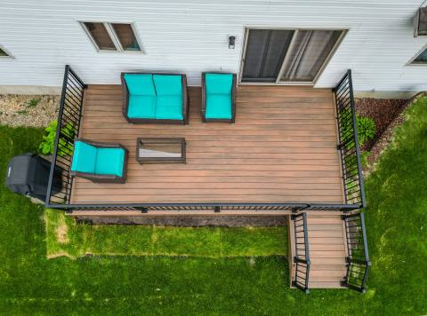 Green Shield Tigerwood deck with black aluminum baluster deck railing. Vibrant deck furniture and table can be seen on a beautiful deck next to a light colored home.