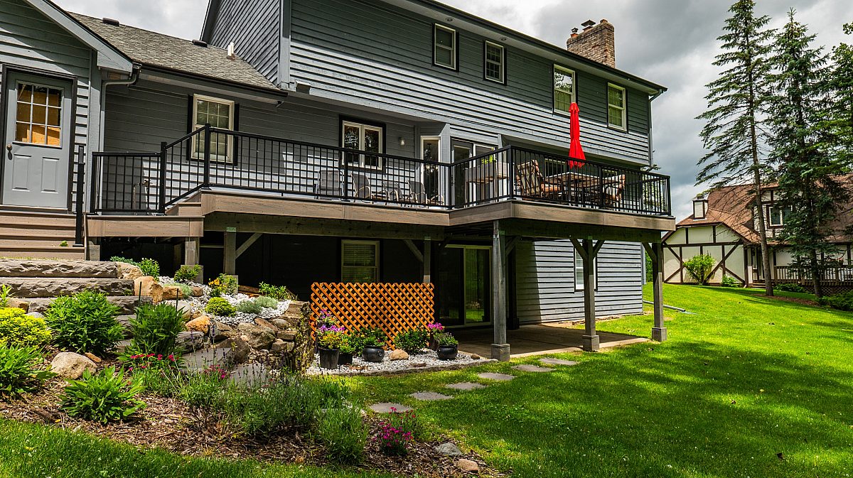 A second-story walkout deck built by Green Shield, featuring Urban Hazelwood composite decking with Amazon Gray accents, black railing, and a view over a grassy, manicured backyard.