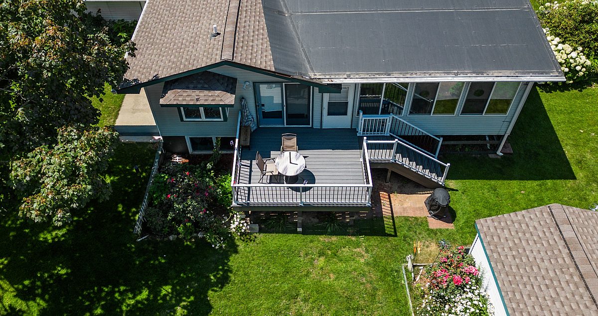 Green Shield deck with white aluminum baluster deck railing, Green Shield standard stairs, and Gray composite next to a light colored house. Drone shot from a high angle, tree tops and flowers can be seen near the deck.